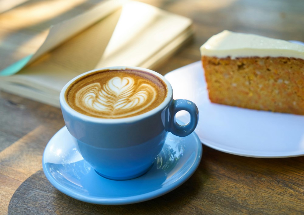 A cup of coffee on a table next to a book and slice of cake to represent a small cafe