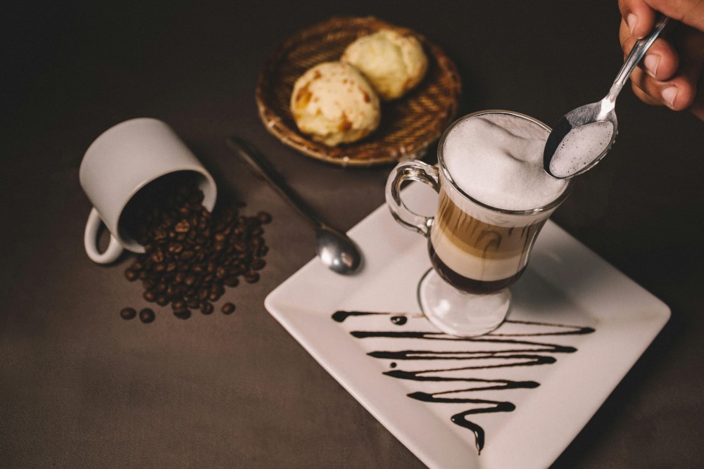 A coffee cup filled with coffee beans and basket with bread on a table behind a tray with a coffe cup and spoon