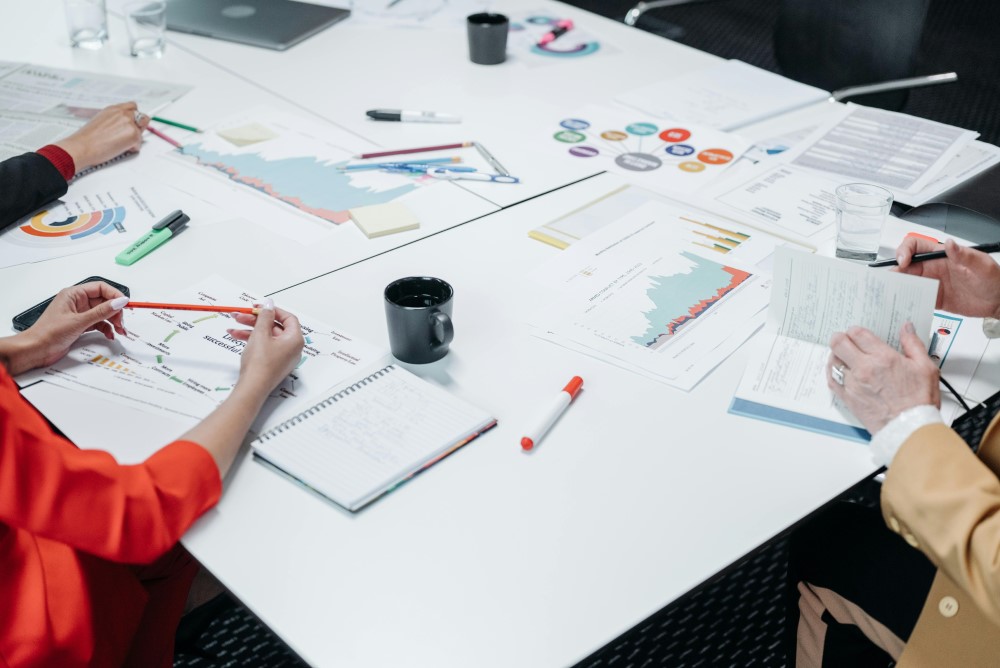 Co-workers surrounding a white desk containing office supplies and various graphs